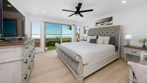 A bedroom with a bed, a television, a ceiling fan, a nightstand, and a view of the ocean.