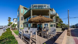 A two-story building with a balcony, outdoor seating area with chairs and a table, and a thatched umbrella.