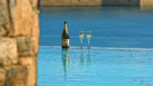 Una botella de champán y dos copas en una piscina con un muro de piedra y vistas a un paisaje montañoso.