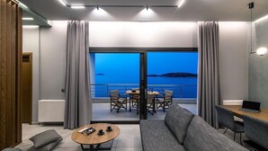 Uma sala de estar moderna com sofá, mesa de centro e área de jantar. Possui uma grande porta de vidro deslizante que dá para uma varanda com vista para o mar.