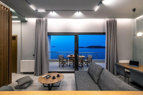 A modern living room with a sofa, coffee table, and dining area. It features a large sliding glass door leading to a balcony with a view of the sea.