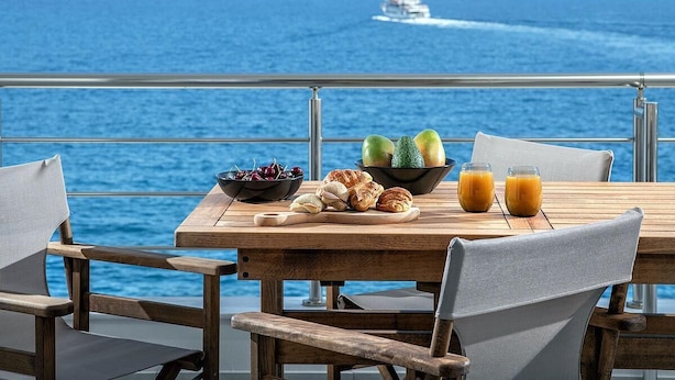 Uma mesa de madeira com uma tigela de frutas, croissants e copos de suco, com vista para o mar e uma ilha ao longe.