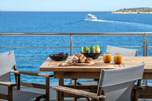 A wooden table with a fruit bowl, croissants, and juice glasses, set against a backdrop of the sea and a distant island.