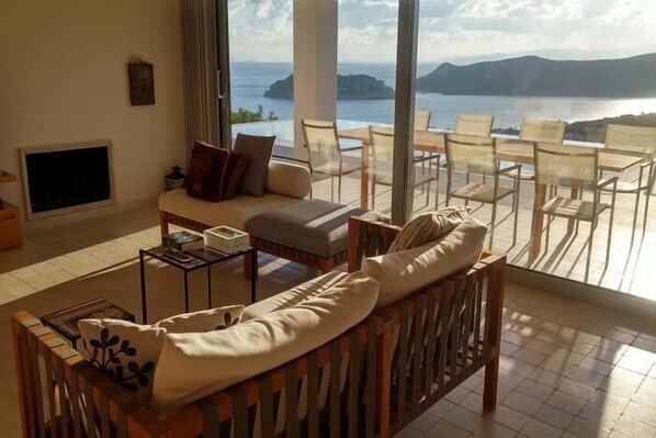 Ein Wohnzimmer mit Sofa, Couchtisch und Fernseher. Ein Balkon mit Tisch und Stühlen bietet Blick auf ein Gewässer und Berge.