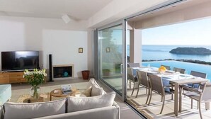 A modern living room with a flat-screen TV, a sofa, and a coffee table. There's a dining area with a table set for four and a view of the sea through large glass doors. - Sea view 3 bedroom villa close to Plaka beach (Elounda)