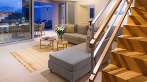 A modern living room with a staircase, a sofa, a coffee table, and a dining area with a table and chairs. - Sea view 3 bedroom villa close to Plaka beach (Elounda)