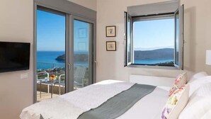 A bedroom with a bed, a TV, a window with a view of the sea, and a balcony with outdoor furniture. - Sea view 3 bedroom villa close to Plaka beach (Elounda)