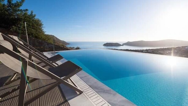A clear blue infinity pool with a wooden deck and lounge chairs, overlooking a calm sea and distant hills. - Sea view 3 bedroom villa close to Plaka beach (Elounda)