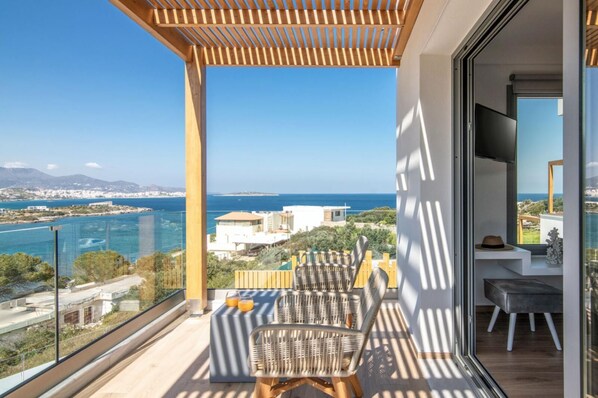 Un balcón con vistas al mar y edificios, una mesa con sillas, y un ventanal de vidrio que da al interior.