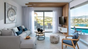 A modern living room with a sofa, ottoman, and chairs, a TV, and a view of a pool and landscape. - Villa Anemos Heated pool (Agios Nikolaos)