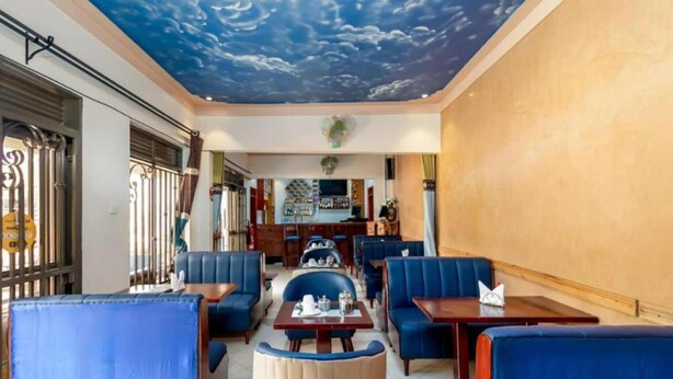 A restaurant with blue upholstered booths and tables, a bar area in the background, and a ceiling with a cloud mural. - Haber Motel  (Entebbe)