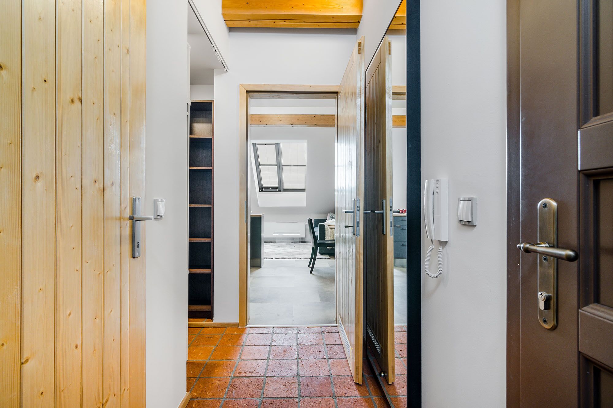 A hallway with a wooden door on the left, a glass door on the right, and a tiled floor. There is a small window with blinds in the background.