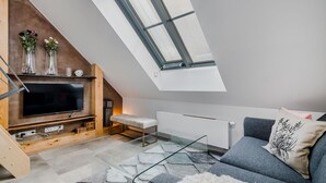 A modern loft bedroom with a sloped ceiling, a large skylight, a flat-screen TV, a glass coffee table, and a grey sofa with a floral pillow.