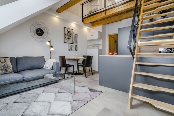 En modern lägenhet i loftstil med en säng på entresolplan, ett matområde och ett vardagsrum med soffa och soffbord.