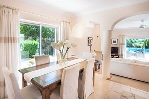 Relax in a spacious living area with a dining table for four, comfortable sofa, and TV, surrounded by lush garden views and a pool.