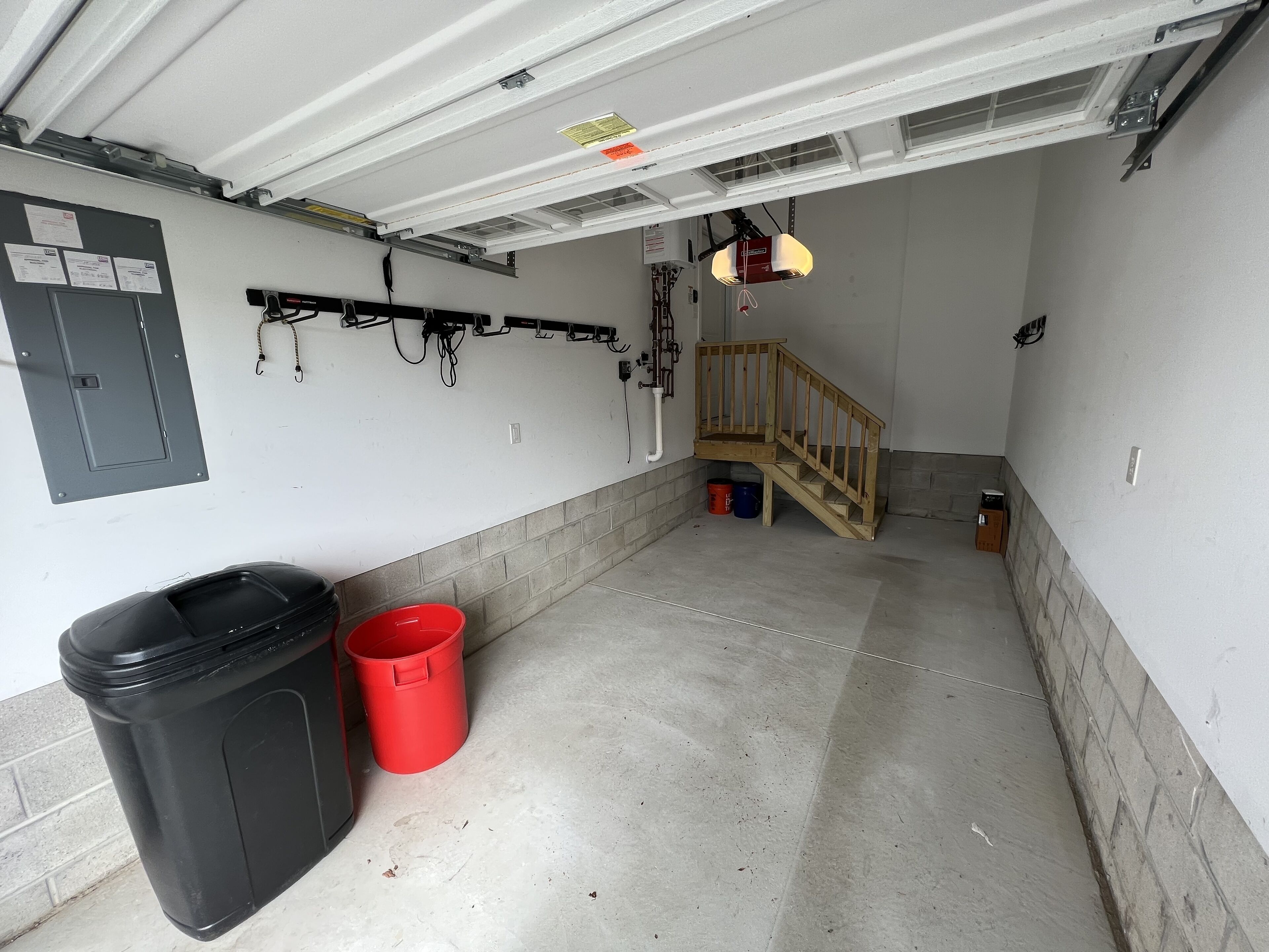 Een garage met een betonnen vloer, een houten trap, een zwarte prullenbak en een rode emmer.