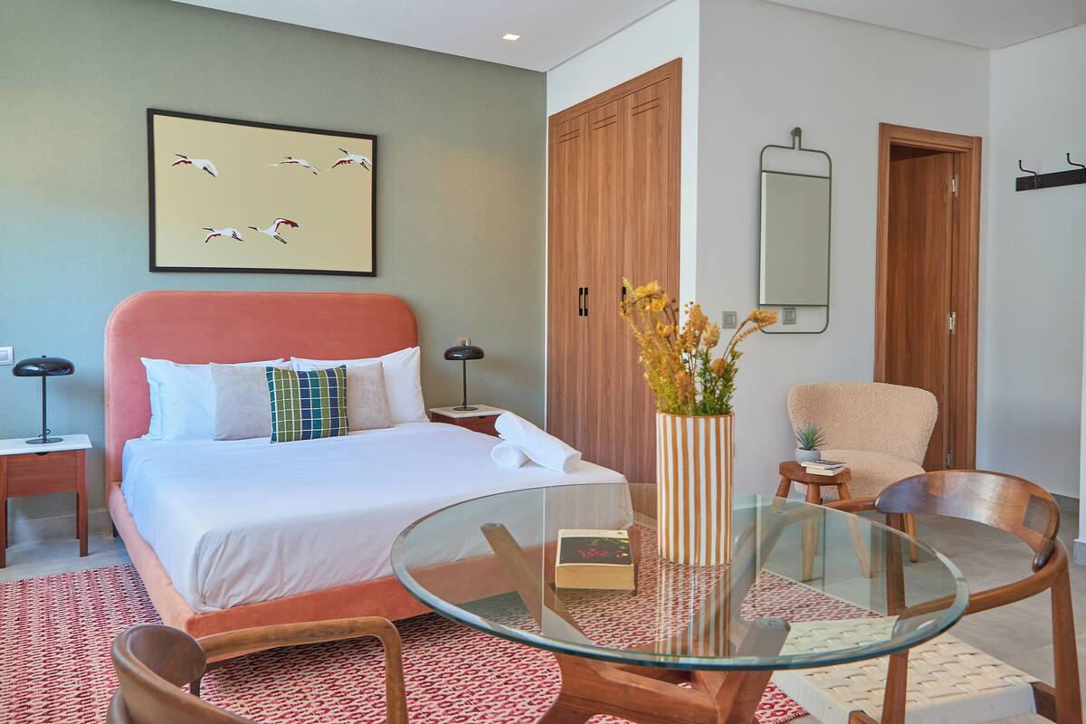 A hotel room with a bed, a glass table, a chair, a small table with a plant, and a mirror on the wall.