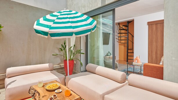 Un área de estar moderna al aire libre con un parasol a rayas, sofás acolchados, una mesa de centro de madera con aperitivos y bebidas, y una planta en maceta.