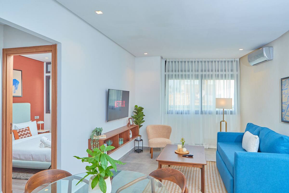 A modern living room with a blue sofa, a wooden coffee table, a glass dining table, and a TV on the wall.