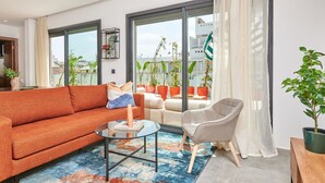 Una sala de estar moderna con sofá, sillón y mesa de centro. Amplias ventanas con cortinas, vistas a edificios exteriores y una planta en maceta a la derecha.