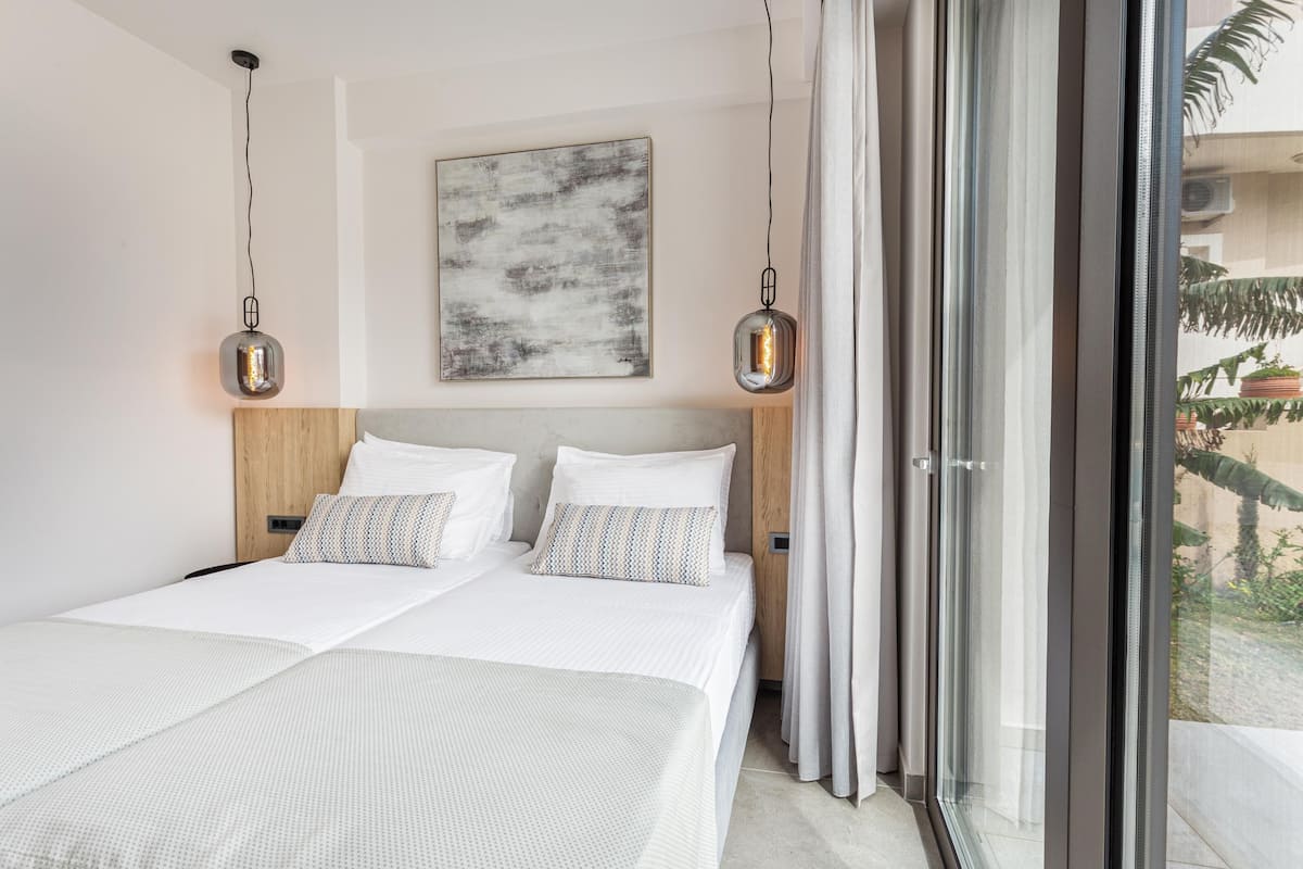 A modern hotel room with a large bed, two bedside tables, a wooden headboard, and a view through a sliding glass door.