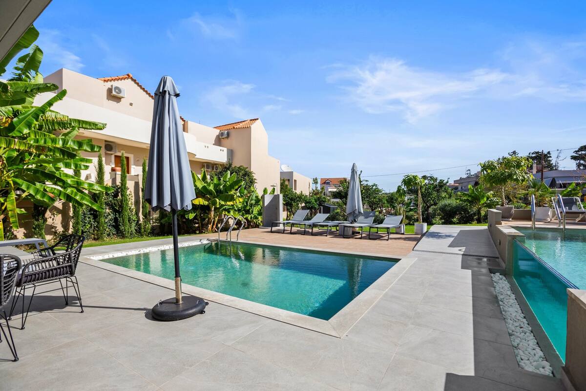 A pool area with lounge chairs and umbrellas, surrounded by greenery and buildings.