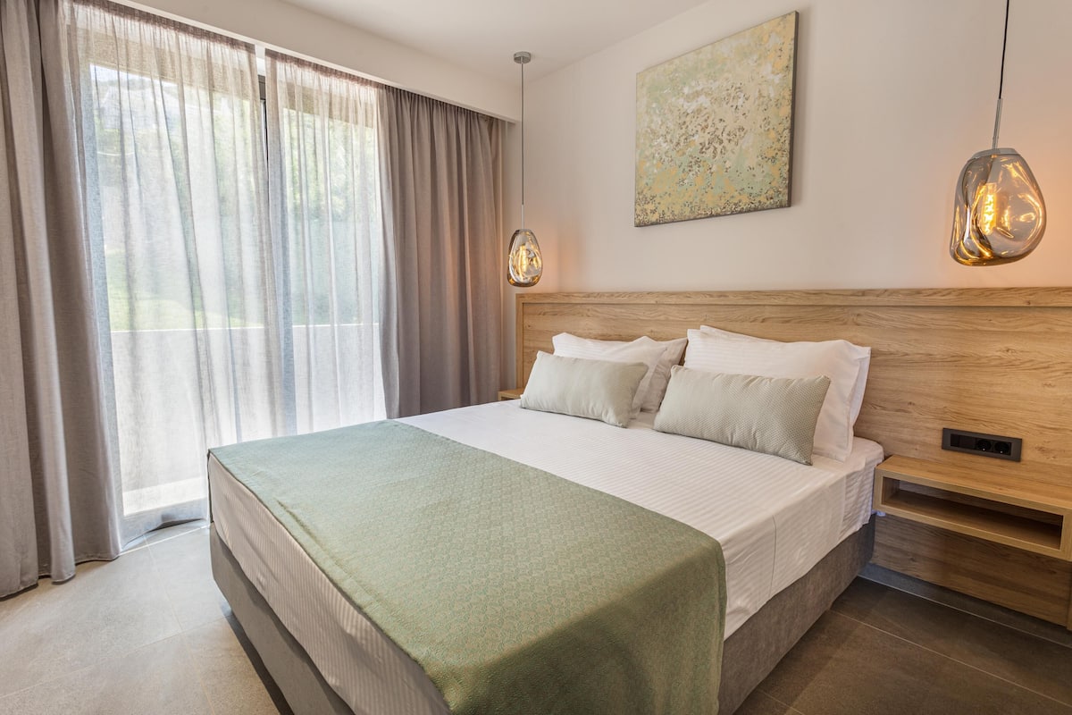A modern hotel room with a large bed, a wooden headboard, a bedside table, and a large window with curtains.