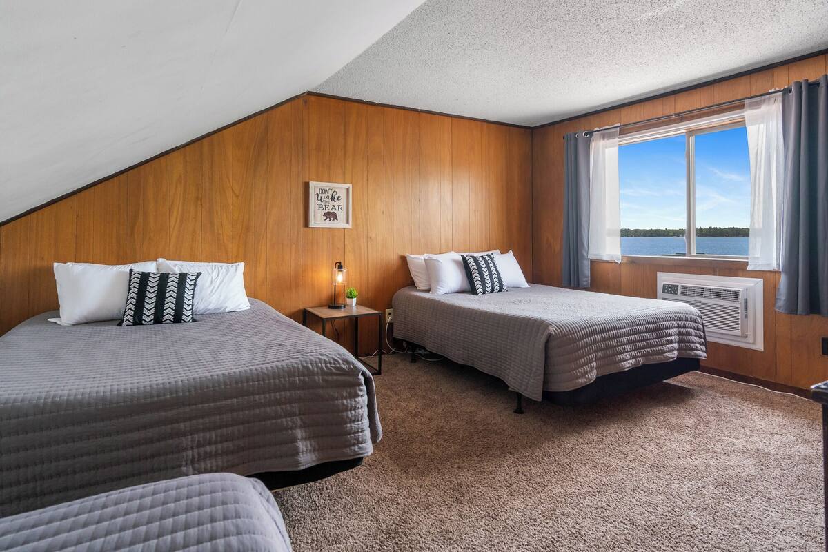 Une chambre avec deux lits, une petite table avec une lampe, un tableau au mur et une fenêtre donnant sur l’eau.