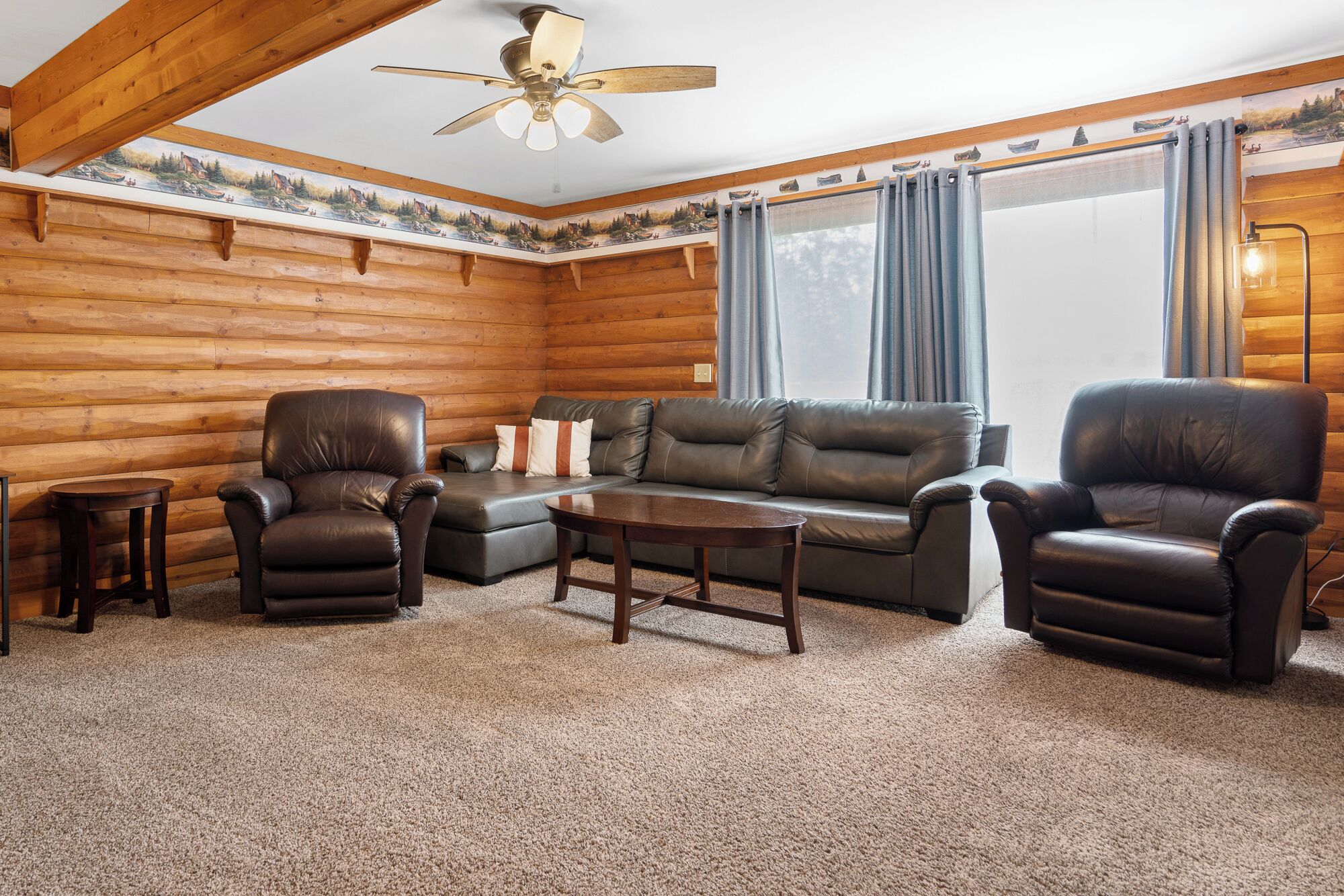 A log cabin interior with a sofa, two leather chairs, a small table, and a ceiling fan.