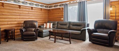 A log cabin interior with a sofa, two leather chairs, a small table, and a ceiling fan.
