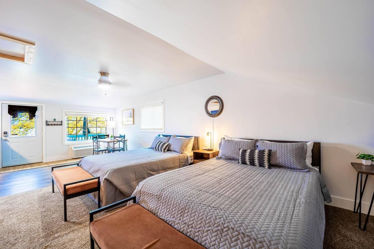 Une chambre avec deux lits, une petite table, un banc, un miroir et une fenêtre avec des stores.