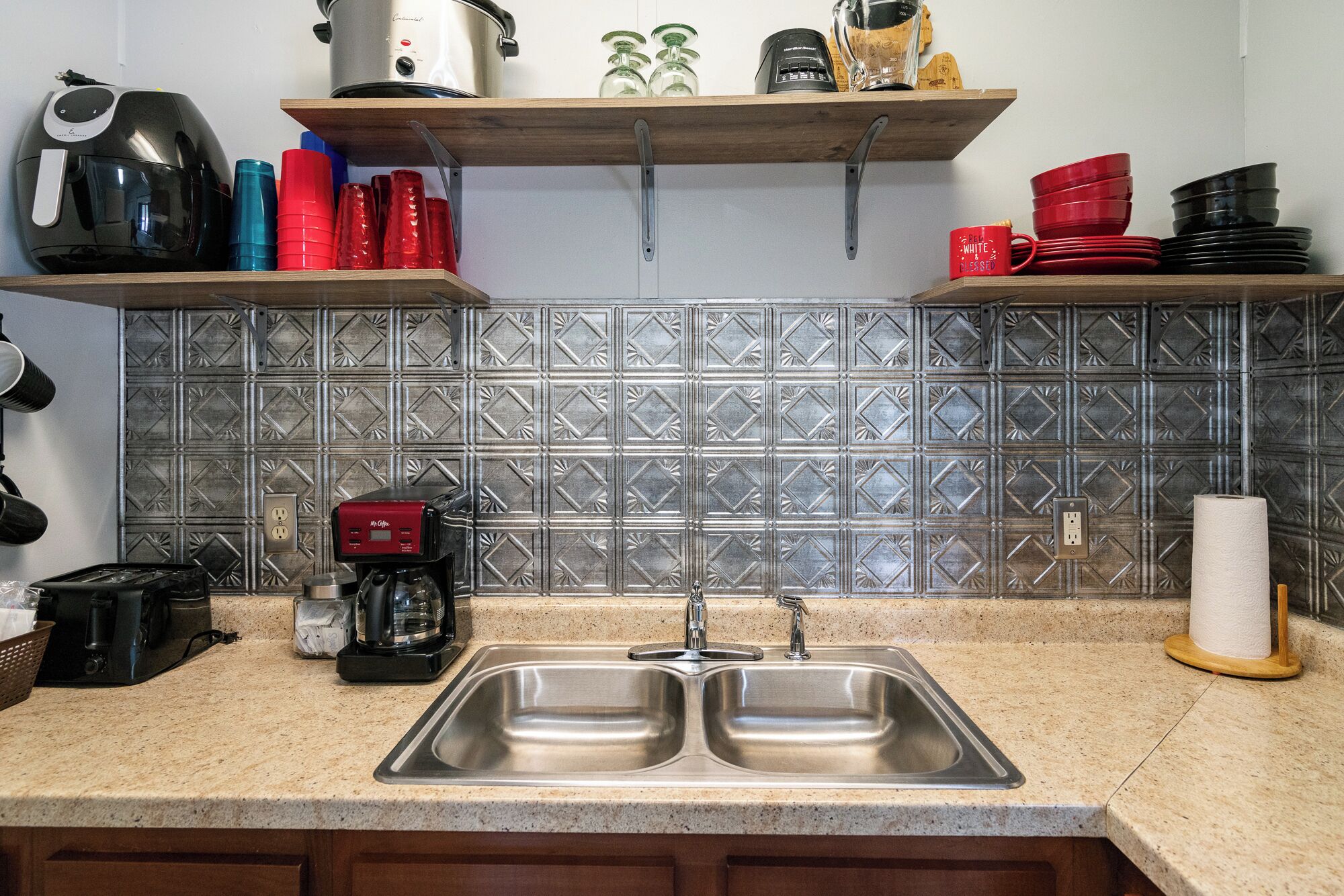 Una cocina con un lavabo doble, una cafetera, un tostador y una tetera sobre la encimera. Hay estantes encima de la encimera con distintos utensilios de cocina.