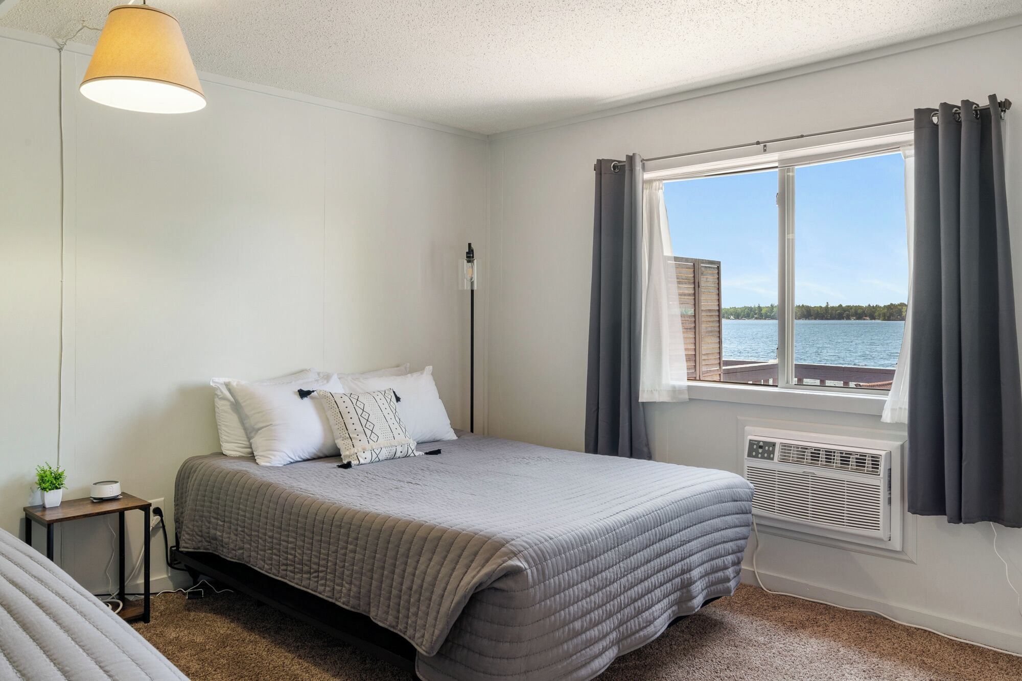 A bedroom with a bed, a bedside table with a lamp, a window with curtains, and a view of a body of water.