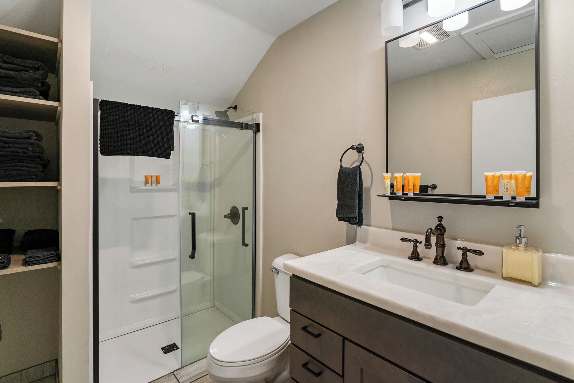 Une salle de bain avec une douche à parois de verre, des toilettes blanches, un meuble-lavabo avec un miroir et des étagères contenant des articles de toilette.