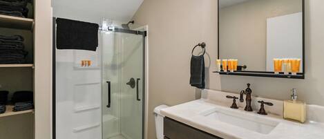 Une salle de bain avec une douche à parois de verre, des toilettes blanches, un meuble-lavabo avec un miroir et des étagères contenant des articles de toilette.