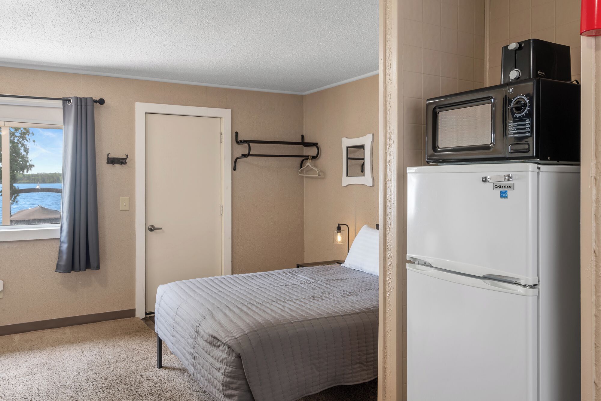 Una habitación pequeña y funcional con cama, refrigerador blanco, microondas, puerta y ventana con cortinas.