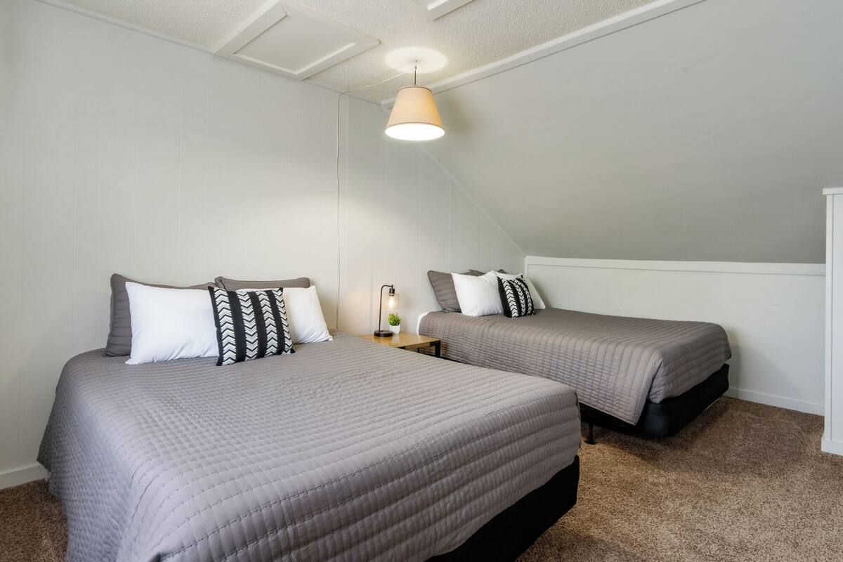 Une chambre à deux lits, chacun étant recouvert d’une couette matelassée grise et agrémenté d’oreillers à motifs. On aperçoit une table de chevet avec une lampe et une petite plante. Les murs de la chambre sont blancs et le plafond est en pente, avec un seul luminaire.