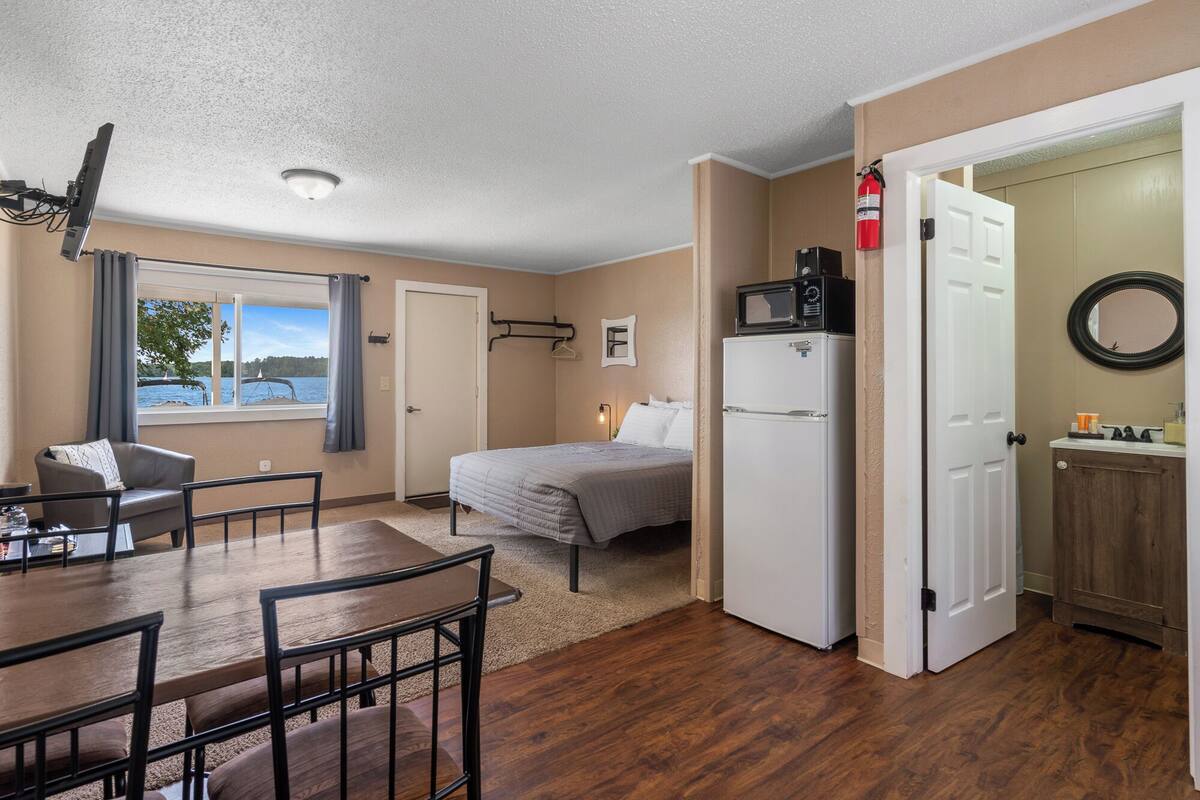 Une pièce compacte comprenant un lit, une table à manger avec des chaises, une petite kitchenette et une vue sur un lac par la fenêtre.