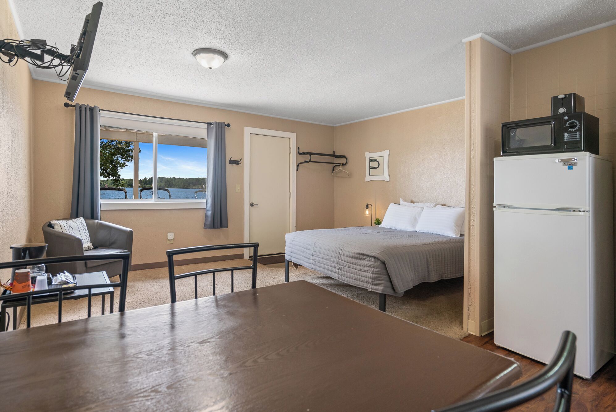 Habitación de hotel con cama, mesa de comedor, una silla, refrigerador, microondas, ventana con vista a un lago e iluminación en el techo.