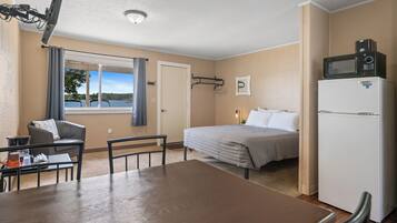 Ett hotellrum med en säng, ett matbord, en stol, ett kylskåp, en mikrovågsugn, ett fönster med utsikt över en sjö och en taklampa.