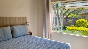 Un dormitorio con una cama grande, cabecera de madera, mesita de noche y ventana con vistas a zonas verdes y una casa.