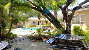 Zona junto a la piscina con una mesa de madera y bancos, rodeada de exuberante vegetación y un edificio al fondo.
