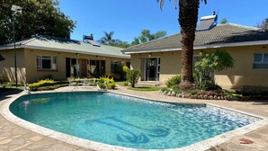 A swimming pool with a stone border, surrounded by a patio area with plants and a house in the background.