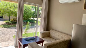 A room with a sofa, a small table, and a refrigerator. There is a sliding glass door leading to an outdoor area with greenery.