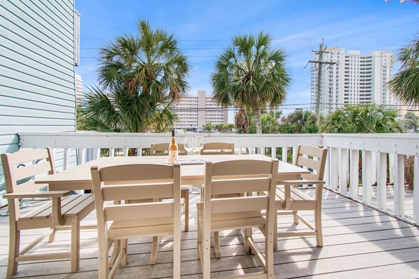 A wooden deck with a table and chairs, surrounded by a white railing, palm trees, and buildings in the background.