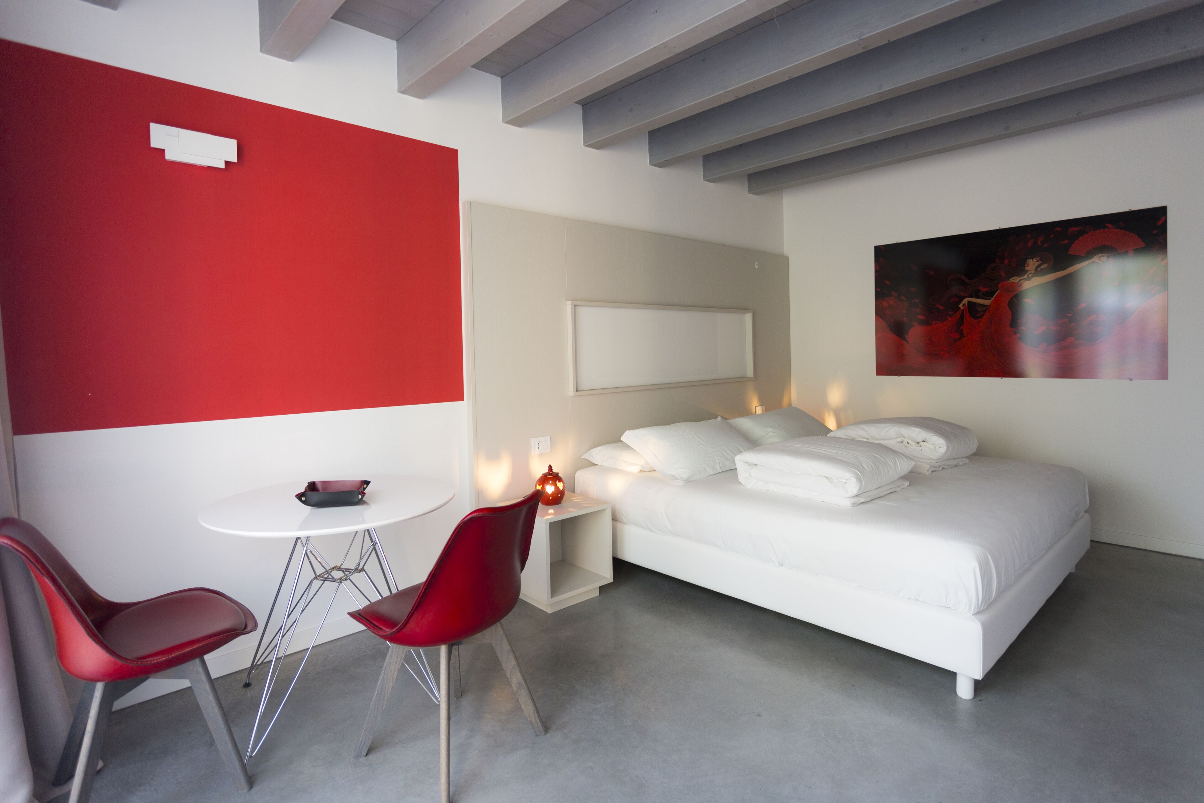 A modern hotel room with a red accent wall, a white bed, a red chair, a small table, and a painting on the wall.