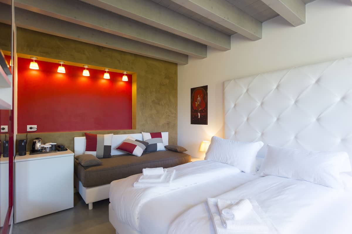 Une chambre d’hôtel moderne avec un grand lit, un canapé, un petit bureau avec une bouilloire et un mur d’accent rouge.
