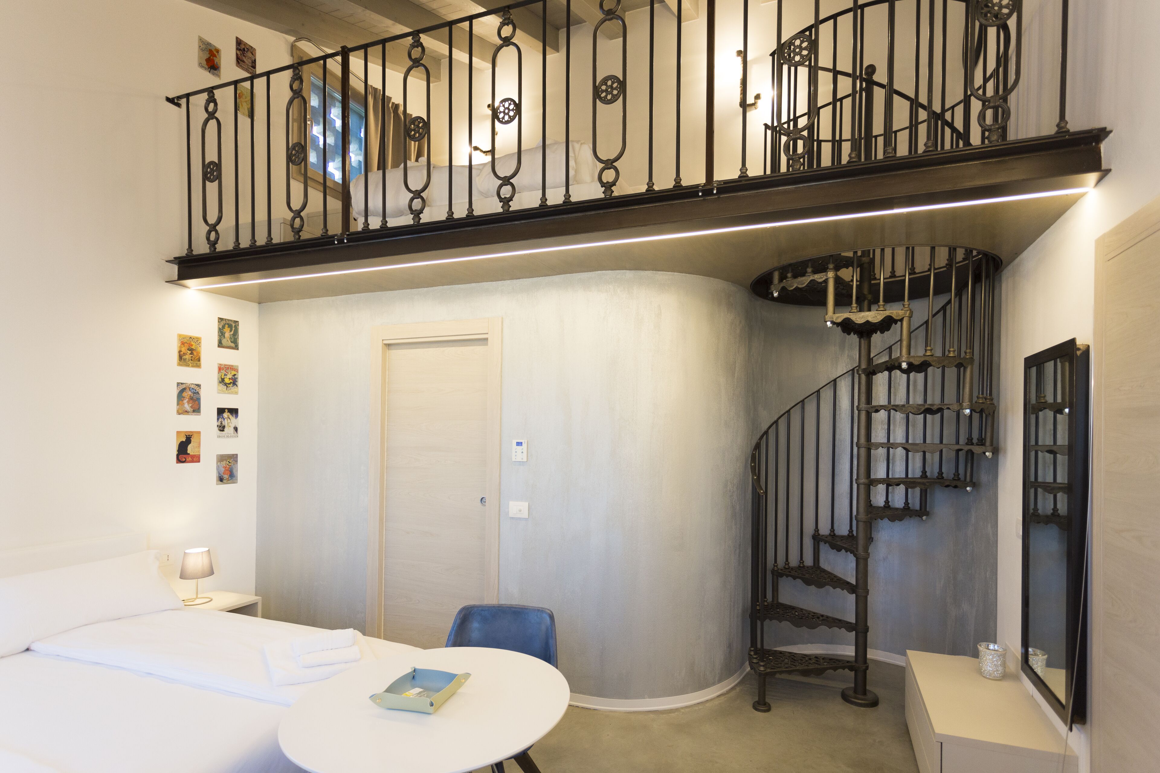 Una habitación de hotel con una cama, una mesa redonda, una silla, un escritorio, una mesita con un florero, un espejo, una puerta, una escalera y una barandilla.