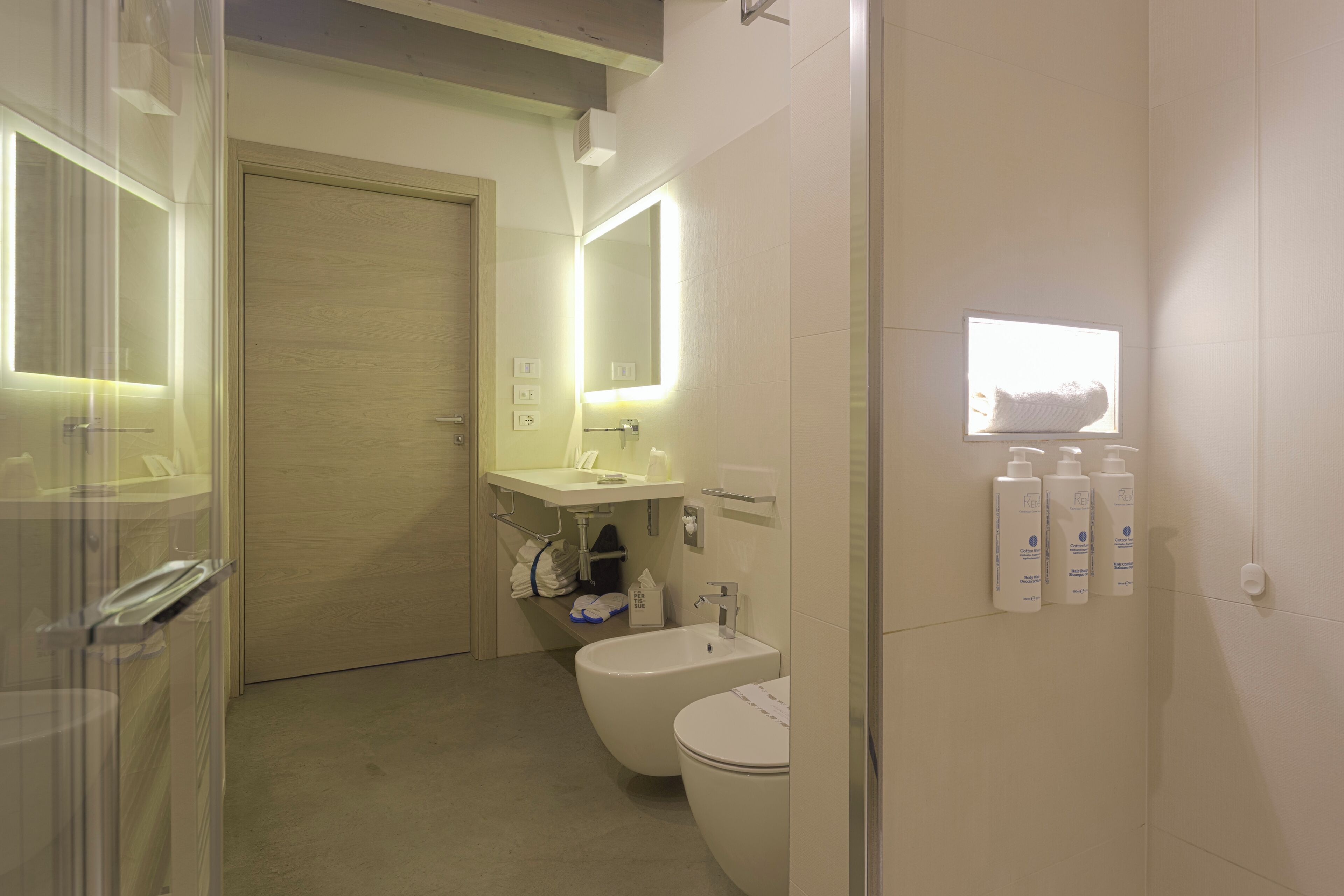 Uma casa de banho moderna com sanita, bidé e lavatório, equipada com espelho e iluminada por luzes de parede.