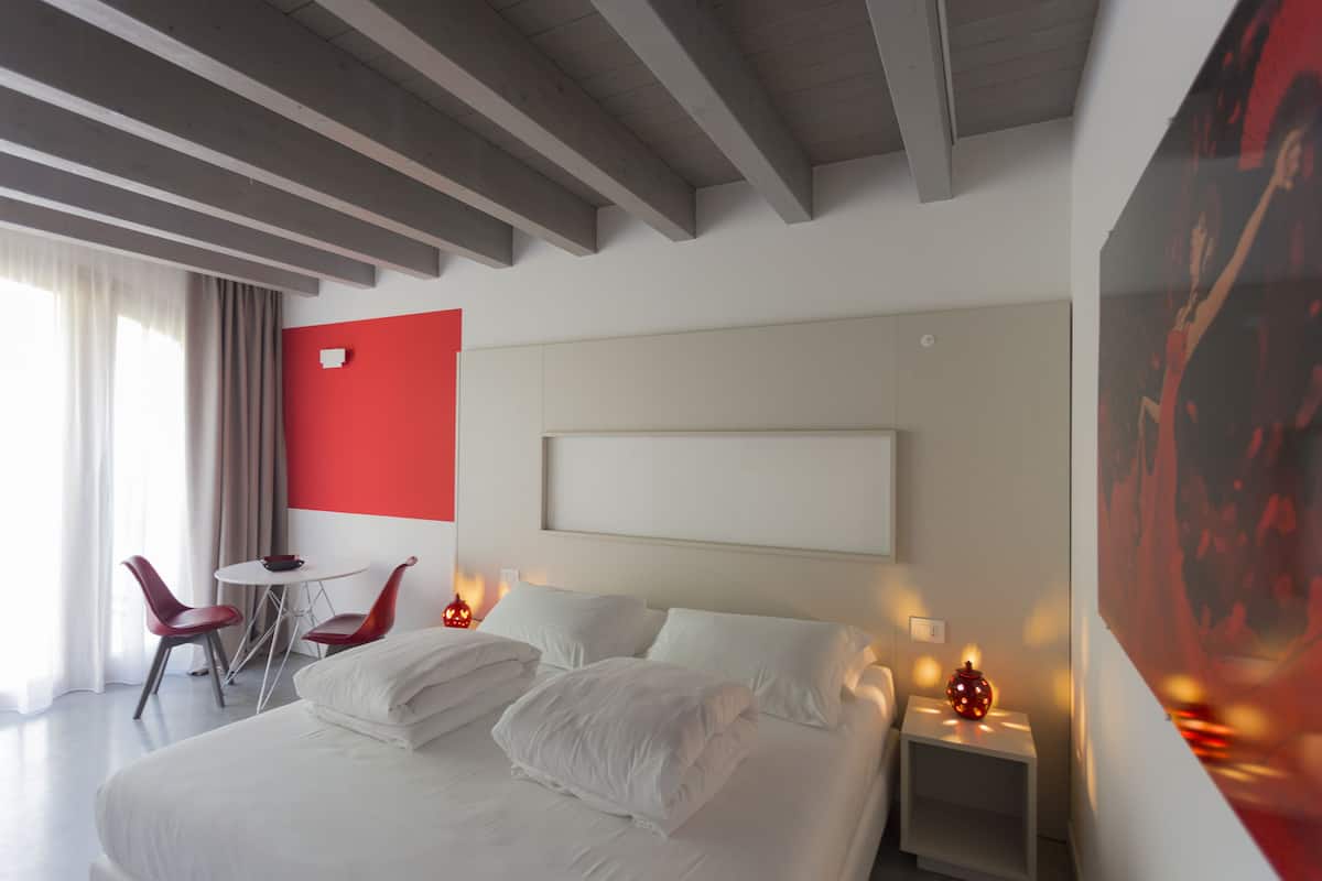 Une chambre d’hôtel avec un lit, deux tables de chevet, un mur d’accent rouge, un plafond blanc et une fenêtre avec des rideaux.